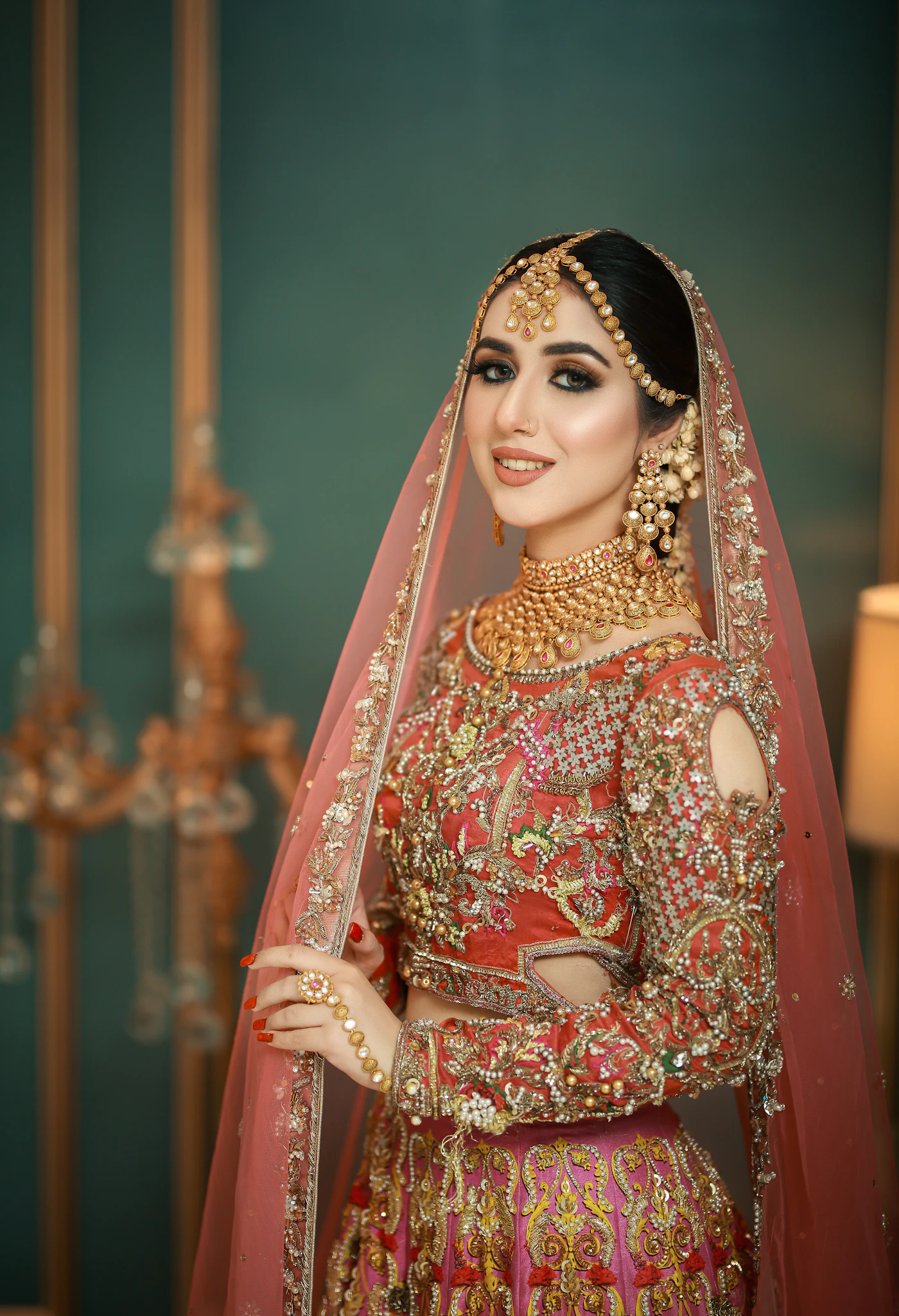 A bride in a red and gold lehenga with a long dupatta, standing in a hall.