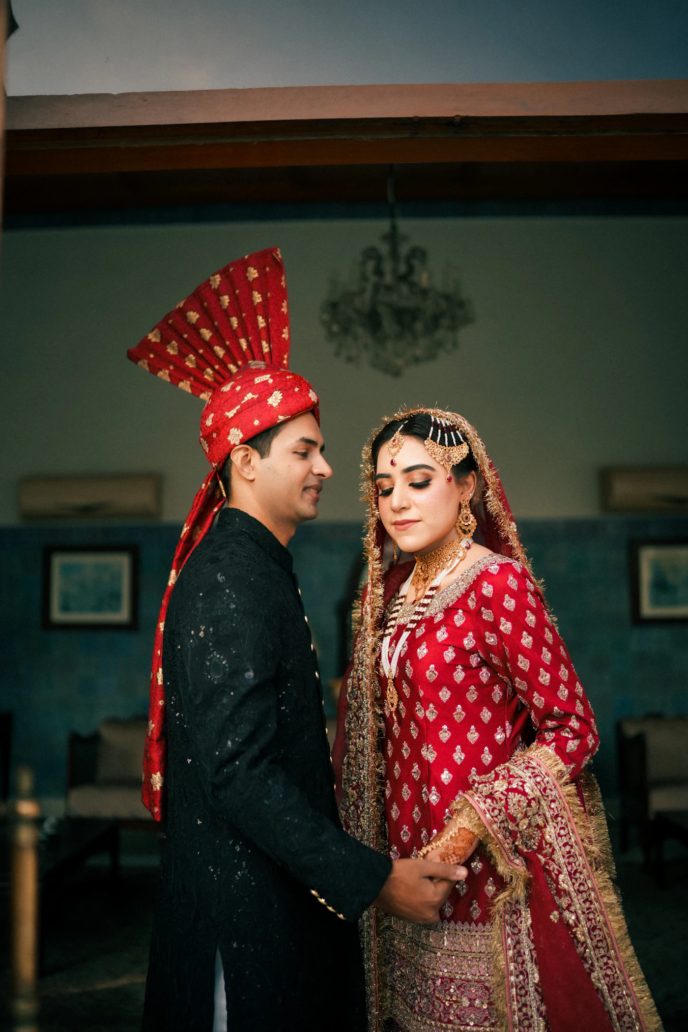 The groom in a turban and the bride in red standing close in a dim room.