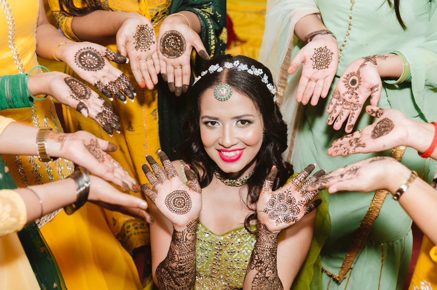 A smiling bride in green with friends holding up their mehndi hands around her.