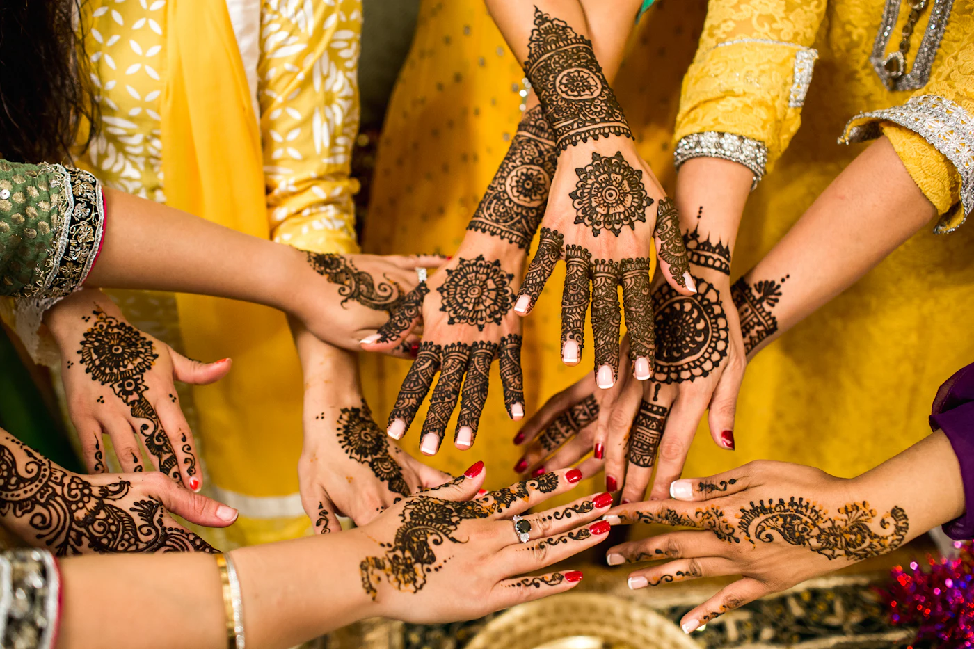 Hands with dark mehndi patterns held together over yellow clothes.