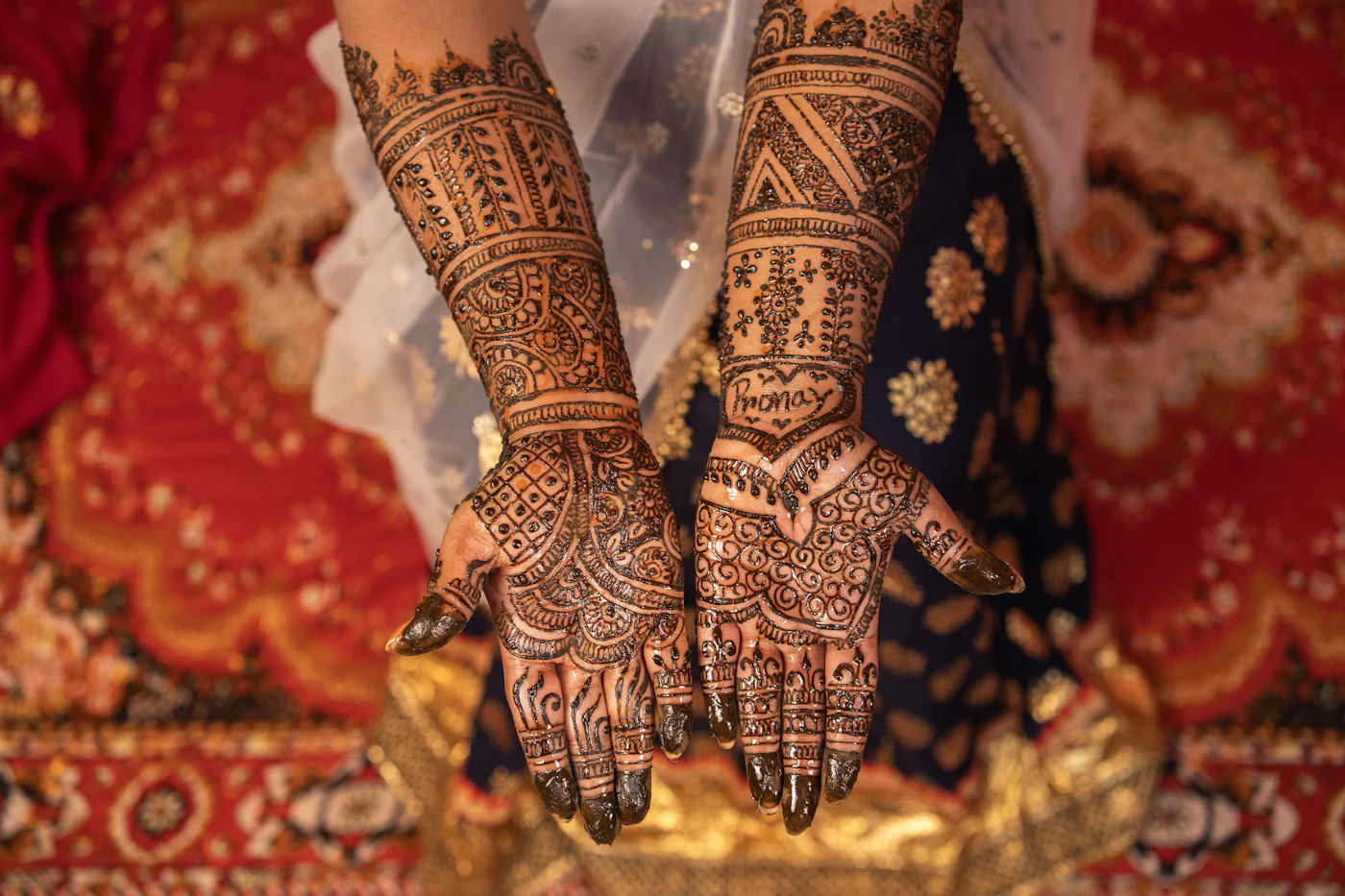 Close up of two hands covered in fine mehndi over a red carpet.