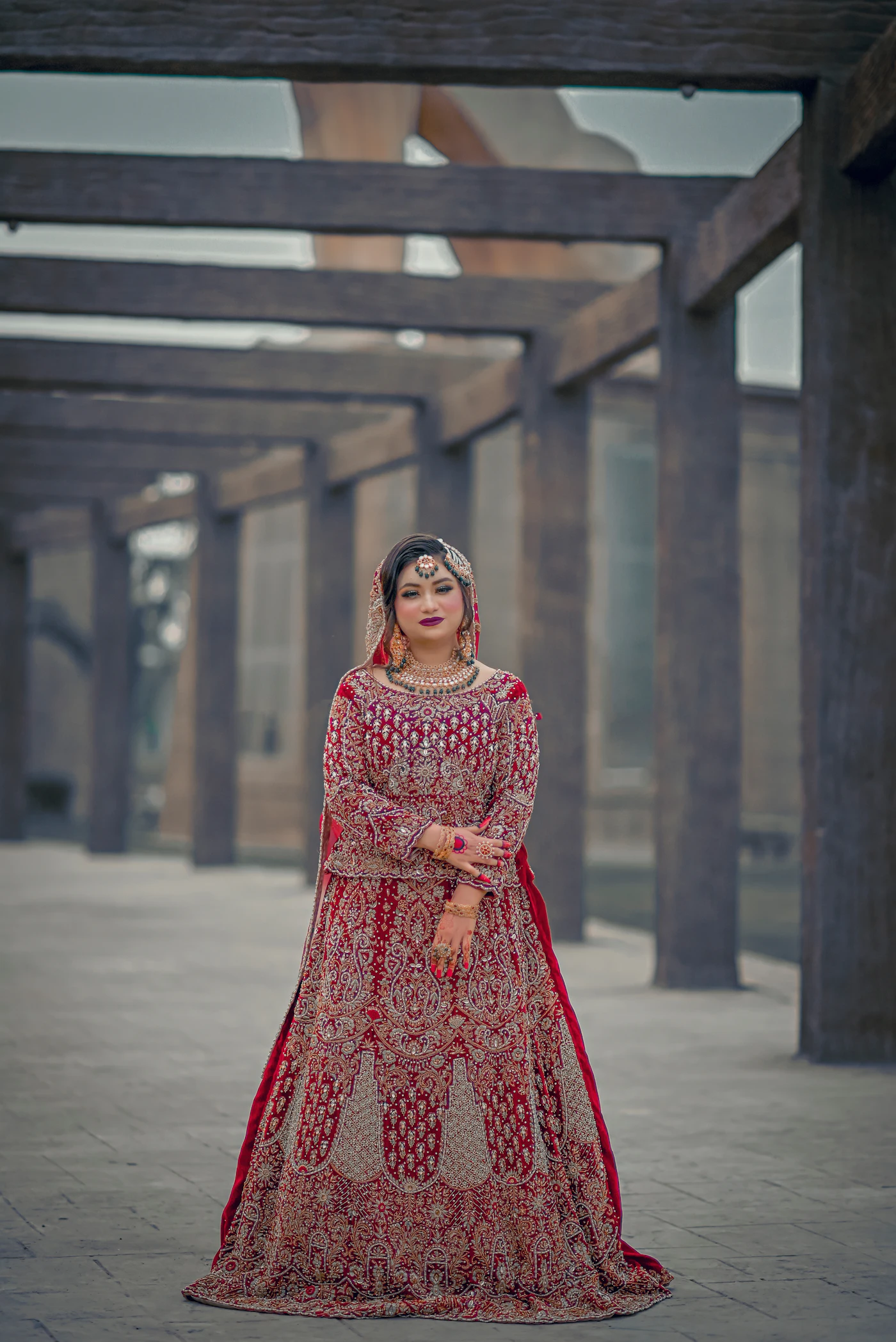 A bride in a deep red gown standing under a wooden pergola.