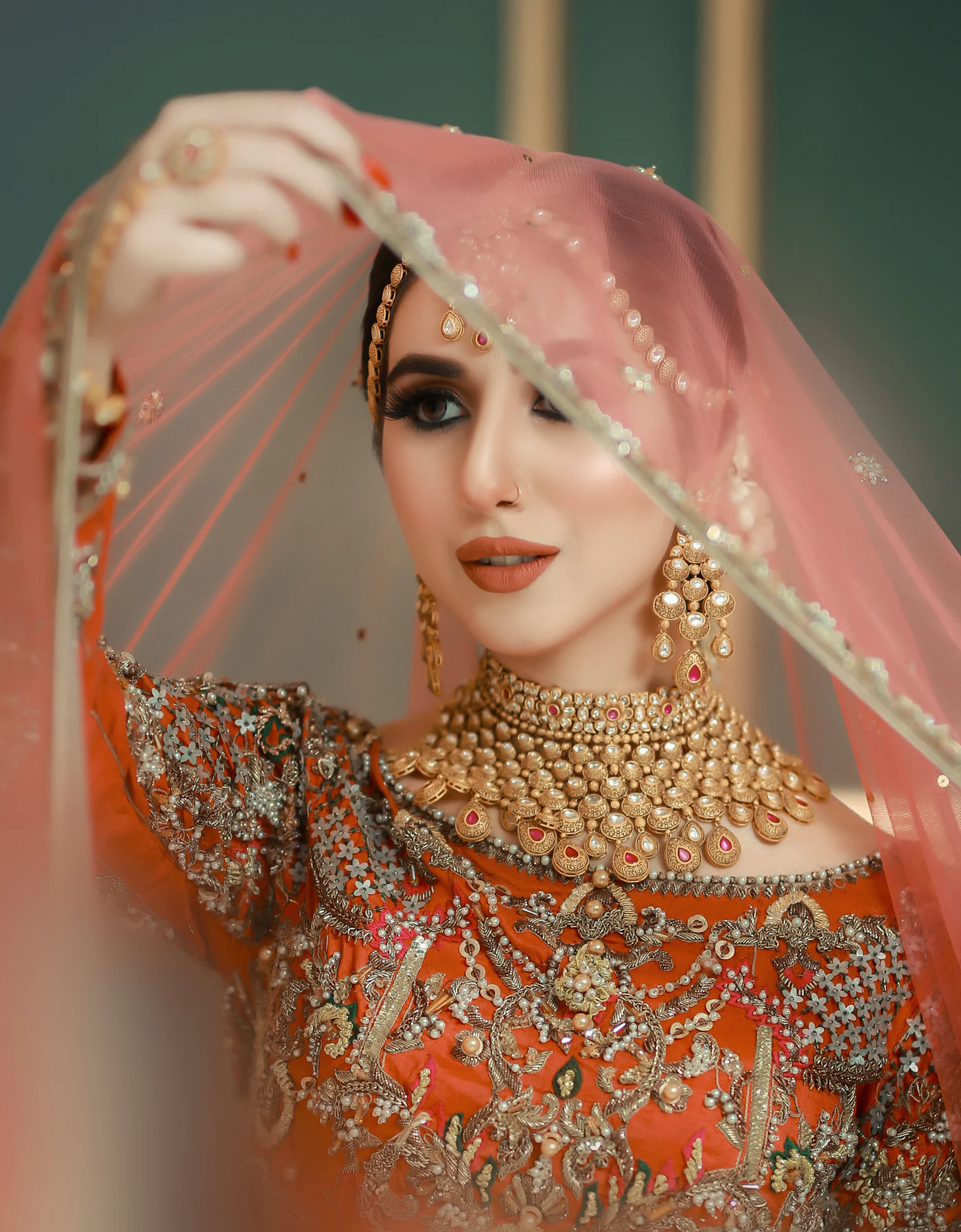 A bride lifting a peach dupatta over her head, wearing a gold necklace.